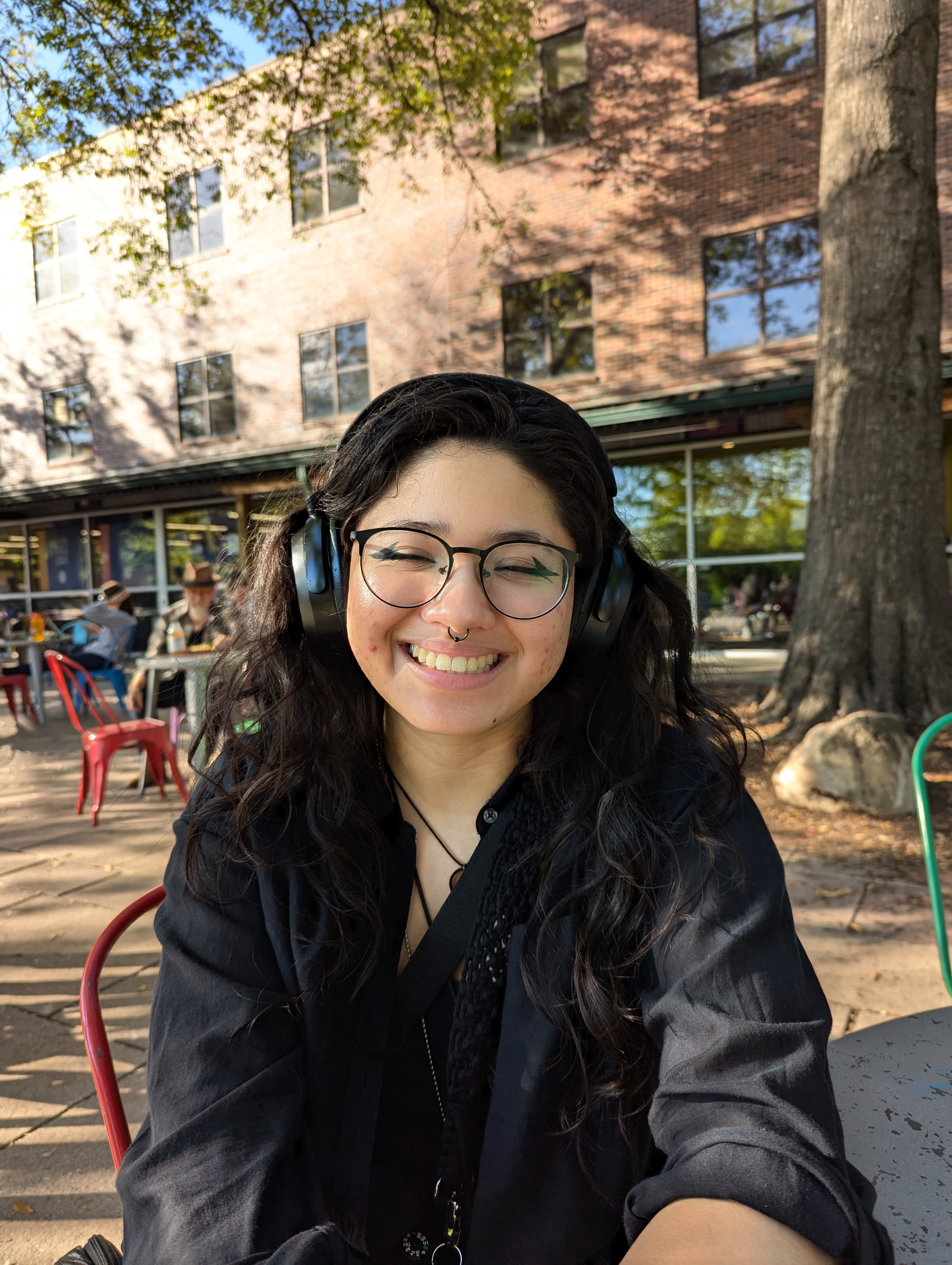 girl with glasses, headphones, long hair and black clothes behind a building and tree.